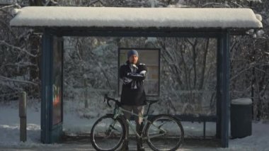 Frozen male cyclist waiting for bus at halt stop with bicycle in winter in snowy weather. Velocipede broke down and man waits long time for transport at the bus stop in winter after a snow storm. 