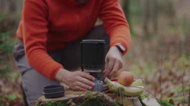 Traveler prepares to eat camping meals and fires up spark button gas stove in portable camping cooking system to boil water in woods in autumn. Inventory for cooking. Hiker igniting camping stove. 