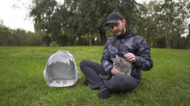 Caucasian man sits on the grass in the park and strokes a pet gray cat, bringing in an animal carrier, a transparent backpack. The theme is a walk with a domestic feline outdoors in cold weather. 