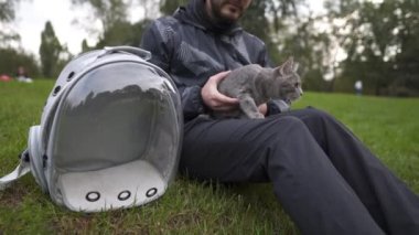 Caucasian man sits on the grass in the park and strokes a pet gray cat, bringing in an animal carrier, a transparent backpack. The theme is a walk with a domestic feline outdoors in cold weather. 