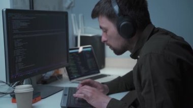 Software engineer wearing headphones is concentrating his work at computer and drinking coffee. Deadlines. Male typing on computer keyboard and developing new program. Coding, developing web site. 