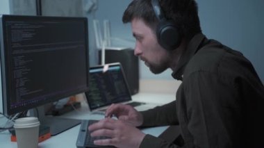 Young male software developer with headsets working, typing on the keyboard and looking focused at the screen. Computer programmer in the headphones is working hard. Developers programming codes. 