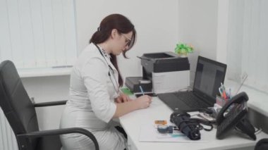 Confident female general practitioner takes notes in medical report book sitting at table in hospital office, professional therapist in white uniform with stethoscope working. Healthcare concept. 
