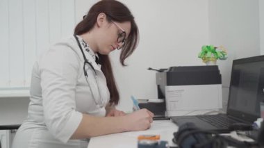 Physician in general practice completing medical card. General practitioner in a white coat does paperwork at the workplace with a laptop, writes notes, prepares documents, reports, prescriptions. 