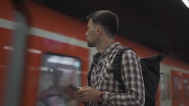 Man with smartphone in front of passing train in Munich, Germany. Passenger with backpack uses phone to create route maps and buy tickets online. S-Bahn Munchen. Muenchen municipal commuter railway. 