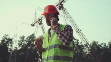 Man working on power line eating sandwich and talking on phone at lunch break. Electrical substation engineer near transmission lines in protective clothing and hard hat having lunch and making call. 