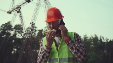 A quick snack on the go. A worker in a hard hat is eating a sandwich and making phone call in front of power line. Electrical engineer having lunch and talking on phone in front of power pole. 