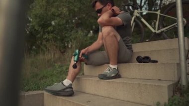 Young male athlete using a massage gun sitting on the outdoor sports ground to warm up and heat up his muscles. Shock pistol after a workout to loosen and relieve pain from muscles. Percussion gun. 