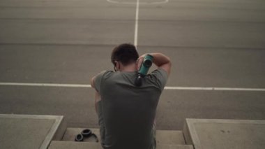 Young male athlete using a massage gun sitting on the outdoor sports ground to warm up and heat up his muscles. Shock pistol after a workout to loosen and relieve pain from muscles. Percussion gun. 