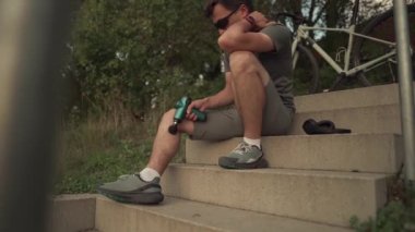 Athlete massages with an impact gun to relieve muscle pain after an outdoor workout. Man massages his body with a percussion pistol after a workout. Warming up muscles with the shock therapy device. 