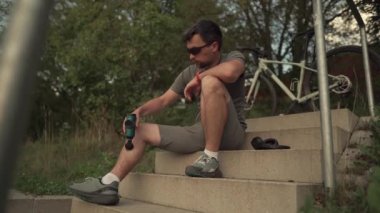 Athlete massages with an impact gun to relieve muscle pain after an outdoor workout. Man massages his body with a percussion pistol after a workout. Warming up muscles with the shock therapy device. 
