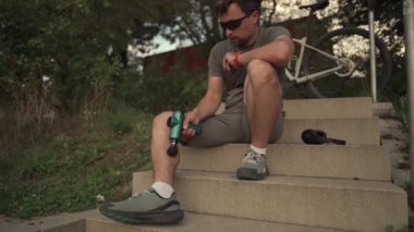 Young male athlete using a massage gun sitting on the outdoor sports ground to warm up and heat up his muscles. Shock pistol after a workout to loosen and relieve pain from muscles. Percussion gun. 