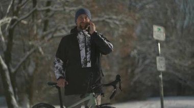 Male cyclist talking on mobile phone standing near bicycle on cycle path in snowy, sunny winter weather in Germany. Cycling in woods in winter. Active lifestyle and commuting on bicycle. ECO friendly