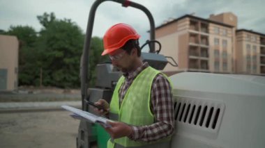 Inspection of civil construction at build site of high rise apartment house. Engineer examines documentation, plan and building progress on drawings in front of machine. Worker against of a paver. 