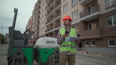 İnşaat alanında kaldırım taşına karşı duran mühendis başlığı teknik belgelerle proje planını kontrol ediyor ve akıllı telefon kullanıyor. İnşaat alanında paten önünde çalışan bir işçi. 