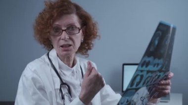 Woman doctor looks into camera and tells patient about results of MRI test. Radiologist explains diagnosis holding CT scan images in hands. Nephropathologist analysis of magnetic resonance imaging. 