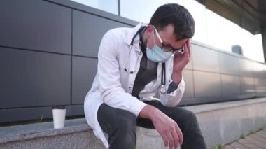 Exhausted young male doctor resting outside hospital drinking coffee to take away. Overtime and overwork in clinic. Healthcare professional takes break. Stressed and tired doctor has cup of coffee. 