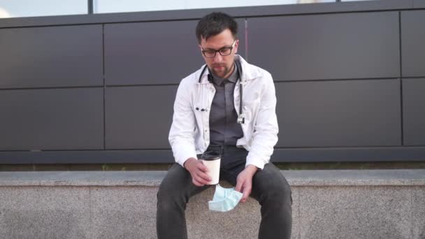 病院の近くの休憩所で疲れた男の若い医者がコーヒーを飲む 若い男性医師は 疲れと落ち着きを感じ 休憩中にコーヒーを飲む 職業の概念 — ストック動画