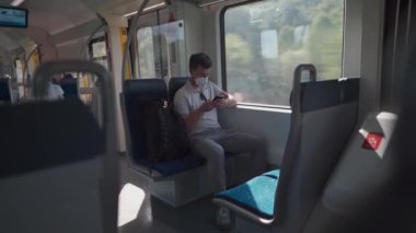 Passenger travelling by train wearing mask, using his phone to browse Internet. Masked man with smartphone on local railway car in Germany. Traveler on railroad with an alergic and air-pollution mask