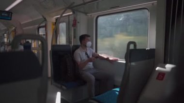Masked man travels on train by window on summer day in sunny weather and uses smartphone. Man traveler in a mask rides a commuter suburban railway in Germany while surfing the Internet on his phone. 