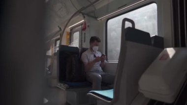 Passenger travelling by train wearing mask, using his phone to browse Internet. Masked man with smartphone on local railway car in Germany. Traveler on railroad with an alergic and air-pollution mask