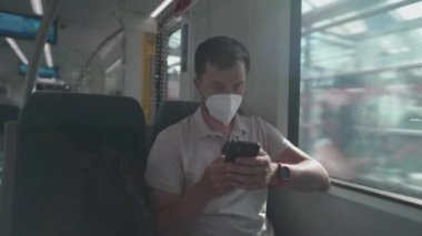 Masked man travels on train by window on summer day in sunny weather and uses smartphone. Man traveler in a mask rides a commuter suburban railway in Germany while surfing the Internet on his phone. 
