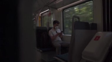 Masked man travels on train by window on summer day in sunny weather and uses smartphone. Man traveler in a mask rides a commuter suburban railway in Germany while surfing the Internet on his phone. 