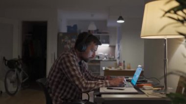 University student learning by laptop with notebook and pen. Online class, E-Learning. Guy wearing headphones typing on keyboard of notebook and making notations in notebook late at night at home. 