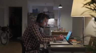 Student sitting at notebook screen with workbook and pen, online lesson, e-learning. Guy with earphones typing on a laptop keyboard and making notes in a sketchbook by pen late at night at home. 