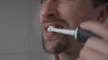 Man brushes his teeth with an electrical tootbrush at morning bathroom before work. Male uses an electric sonic dental cleaning brush. Daily dental hygiene and oral health with eletrical teethbrush. 