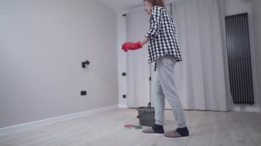 Cheerful mature female cleaning a new apartment without furniture, mopping the floor with a mop dances and sings. Be creative about cleaning your home. People, housework and housekeeping concept. 