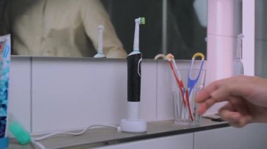Man removes electric toothbrush from charge and brushes teeth before work in morning. Male in shirt picks up the ultrasonic dental brush view of reflection in mirror. Dental cleaner power tool. 