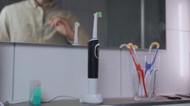 Man removes electric toothbrush from charge and brushes teeth before work in morning. Male in shirt picks up the ultrasonic dental brush view of reflection in mirror. Dental cleaner power tool. 