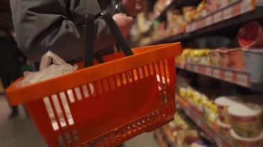 Man with orange grocery shopping cart selecting items at supermarket. Shopping cart holding male while selecting products. Focus on basket of purchases in hand of buyer in store. Choosing products. 