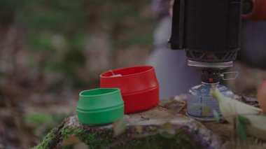 Man hiker making tea while camping, boiling water on burner in woods on stump in autumn. Camping equipment for cooking food and hot drinks. Traveler prepares tea by using system for cooking on gas. 
