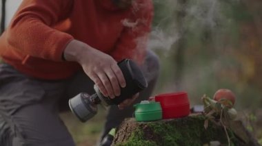 Backpacker cooks tea with integrated system pot and burner on gas fuel cartridge in a forest on stump during a backpacking trip in the fall. Hiker prepare hot drink with portable gas stove. 
