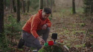 Man hiker making tea while camping, boiling water on burner in woods on stump in autumn. Camping equipment for cooking food and hot drinks. Traveler prepares tea by using system for cooking on gas. 