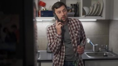 Drunken, irritated man stands with an unfinished bottle of alkogol in hand and calls on phone at his home in kitchen late at night. Alcoholism and depression concept. Drunkenness. Social problems. 