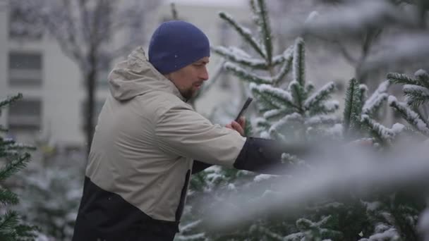 冬の雪の日にクリスマスマーケットでモミの木を選択することについてのオンラインストリーミングサービスでソーシャルメディアネットワークやフォロワーのためのカメラ電話を使用して男性のウェブロガーの撮影ビデオ — ストック動画