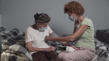 Koronavirüs salgını ve grip sırasında yaşlı bir kadının tansiyonunu ölçen maskeli olgun bir kadın. Bakıcı hasta için elektronik sphygmomanometre kullanıyor. Kan basıncı göstergesi.