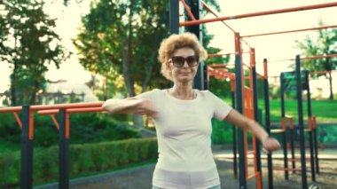 Olgun bir kadın yazın spor salonunda antrenmandan önce ısınıyor. Yaşlı kadın sokak fitness alanında kollarını ve üst omuz bölgesini ısıtmak için egzersiz yapıyor. Emeklilikte sağlıklı yaşam tarzı. 