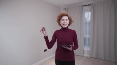 Yaşlı kadın emlakçı yeni dairenin anahtarlarını tutuyor. Olgun kadın emlakçı evin anahtarını verirken arka planda boş, küçük, rahat bir dairede kira için duruyor. Emlak. 