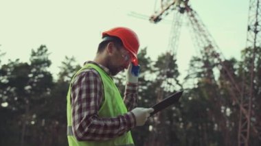 Sert şapka ve güvenlik yeleği giyen elektrik mühendisi, elektrik santralinin yüksek voltajlı elektrik hatlarının yakınında dokümanları olan bir panoyla çalışıyor. Sanayi mühendisi, elektrik hatları ve kuleler. Denetleme