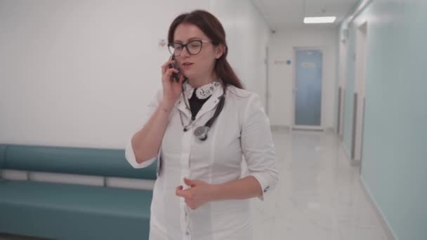 若い女性のドッコールは病院の廊下を歩き 携帯電話で話します 女医は診療所の廊下を電話で通っている 患者と話す医療専門家 — ストック動画
