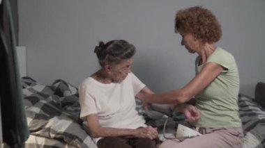 Olgun bir kadın, yaşlı bir kadının kan basıncını sphygmomanometre ile ölçmesine yardım eder. Yaşlı kız hasta yaşlı anneye bakıyor. Hemşire muayenesi hastalığı hipertansiyon.