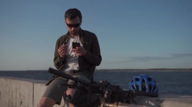 Bisikletçi adam çerez yiyor ve su deposunun arka planında akıllı telefondan internette sörf yapıyor. Erkek, gölün önünde bisiklet sürerken yemek yer ve telefonunu kullanır. Dışarıda bisikletle öğle yemeği yiyorum.. 
