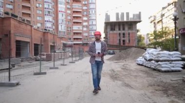 İnşaat mühendisi inşaat alanında yürüyor ve kahve içiyor. Kasketli, kareli gömlekli ve kot pantolonlu bir adam inşaat sahaları boyunca yürüyor ve bir fincan kahve içiyor. Mimarın mola vermesi. 