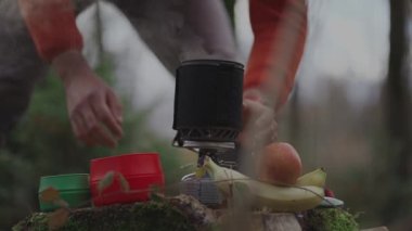 Man hiker making tea while camping, boiling water on burner in woods on stump in autumn. Camping equipment for cooking food and hot drinks. Traveler prepares tea by using system for cooking on gas