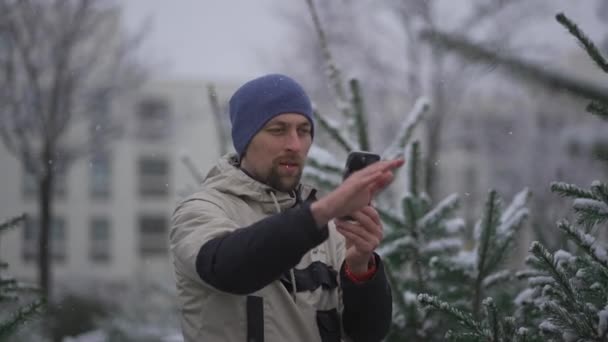 冬の雪の日にクリスマスマーケットでモミの木を選択することについてのオンラインストリーミングサービスでソーシャルメディアネットワークやフォロワーのためのカメラ電話を使用して男性のウェブロガーの撮影ビデオ — ストック動画