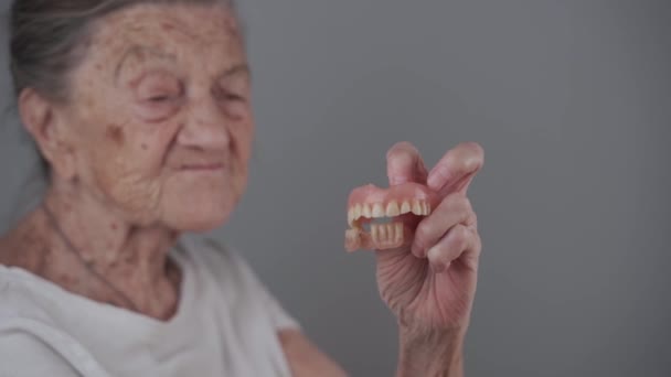 老年妇女得到了可移动的假牙 显示在相机上 口腔护理的概念 假牙和老年 工作室灰白背景的白发老妇人手中的可拆开的完整假肢 — 图库视频影像